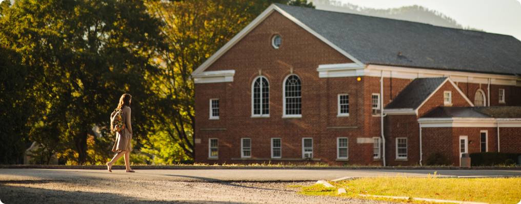 A person carrying a backpack walks across a street toward a large red-brick building at SVU, surrounded by trees on a sunny day—perhaps heading to the college admissions office for financial aid information.