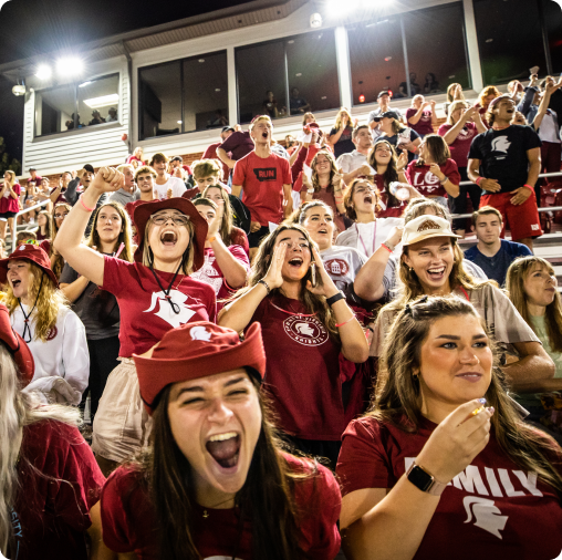 A crowd of enthusiastic fans wearing matching red and white shirts and hats, including many SVU students celebrating their college admissions and financial aid awards, cheer loudly in stadium bleachers during a sporting event.