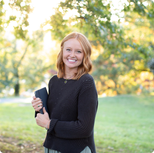 A woman with shoulder-length red hair stands outdoors, smiling and holding a closed laptop or book, with green trees and grass in the background.
