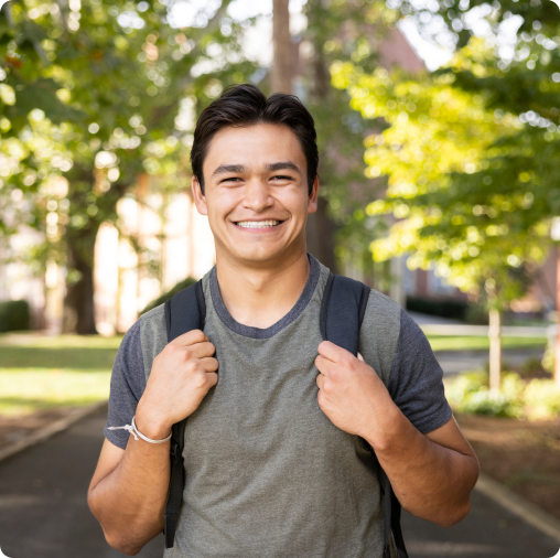 Young man smiling outdoors, wearing a backpack and casual clothes, stands on a tree-lined path—ready for college admissions season or exploring financial aid options at SVU.