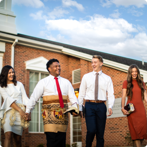 Four young adults in formal attire, holding books, walk and smile together outside a brick church building under a blue sky, enjoying life at SVU after navigating college admissions.