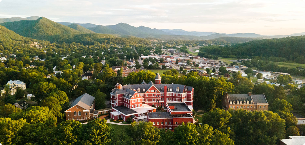 Aerial view of SVU Campus surrounded by trees, with a small town and mountains in the background under a partly cloudy sky.