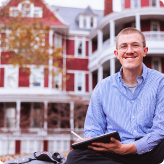 A man wearing glasses and a blue shirt sits outdoors in front of a red brick building, smiling and writing in a notebook, perhaps jotting down thoughts about college admissions or financial aid at SVU.