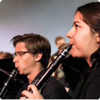 Two musicians play clarinets in an indoor setting, with a blurred background suggesting they are part of a larger college admissions ensemble or orchestra.