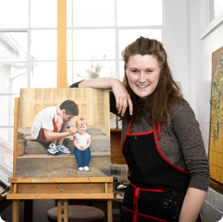 A woman in an art studio stands beside an easel displaying a painting of a man and a child, perhaps discussing college admissions or financial aid as they sit on outdoor steps.