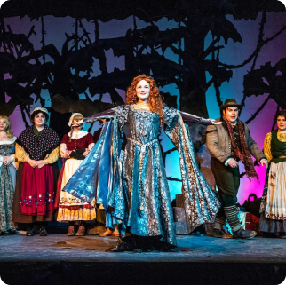 Actors in colorful period costumes perform on stage, with a woman in a blue gown center and others behind her—much like an SVU cast gathering for a dramatic college admissions or financial aid scene against a theatrical backdrop.