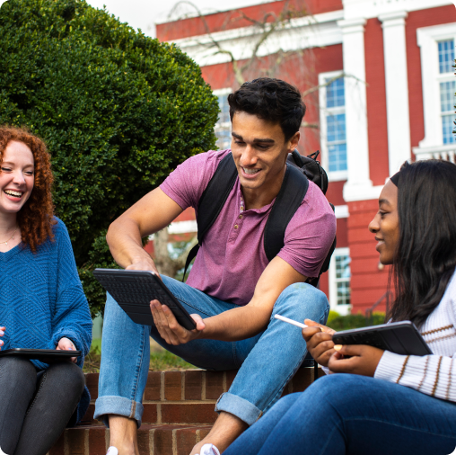 Three students sit outside a red brick building at SVU, smiling and looking at a tablet and notebooks as they discuss college admissions.