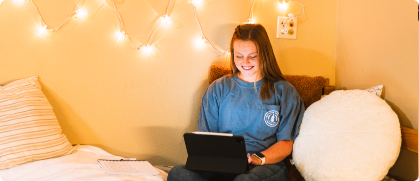A person sits on a bed with string lights on the wall, smiling while using a tablet to check college admissions updates. A notebook and pillow are nearby.